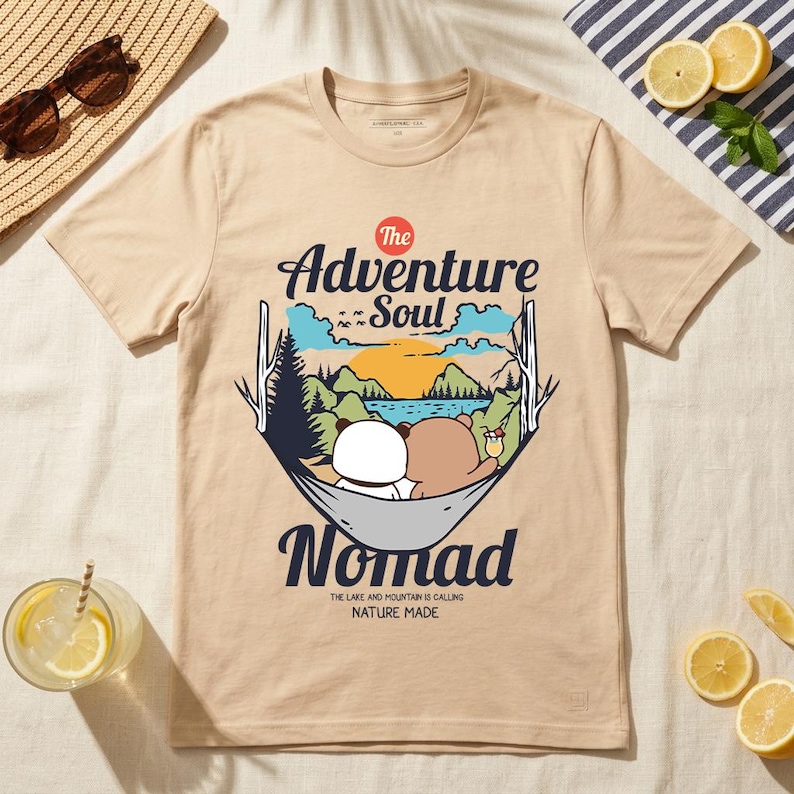 Peut inclure: T-shirt beige avec un motif de deux ours dans un hamac, avec le texte "The Adventure Soul Nomad". Le motif comprend un lac, des montagnes et un coucher de soleil. La phrase "The lake and mountain is calling nature made" est &eacute;galement imprim&eacute;e.