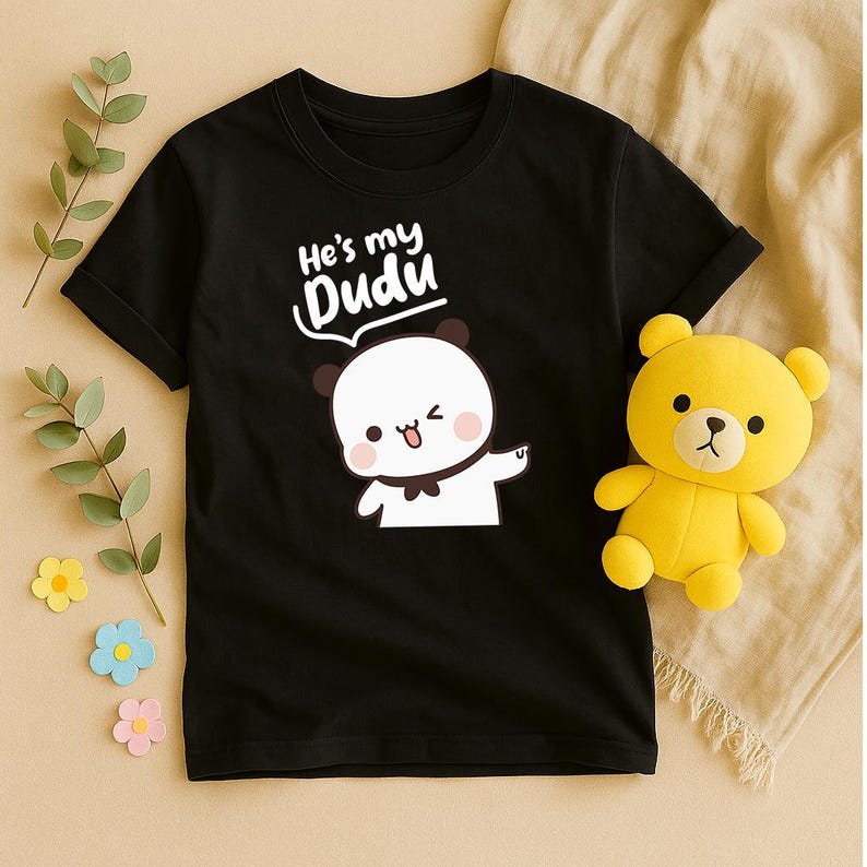 May include: Black t-shirt featuring a cartoon panda with the text "He's my Dudu". A yellow teddy bear is next to the shirt. The shirt has a round neck and short sleeves. The panda has a brown bow tie and is winking.