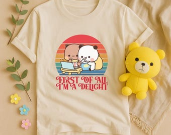 May include: Cream-colored t-shirt featuring a graphic of two cartoon bears with a laptop and food, set against a sunset design. The text "FIRST OF ALL I'M A DELIGHT" is below the graphic. A yellow teddy bear is on the right.