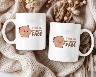 May include: Two white ceramic mugs with a cartoon bear graphic and the text "THIS IS MY HAPPY FACE" in brown. The mugs have a curved handle and are set against a neutral background.