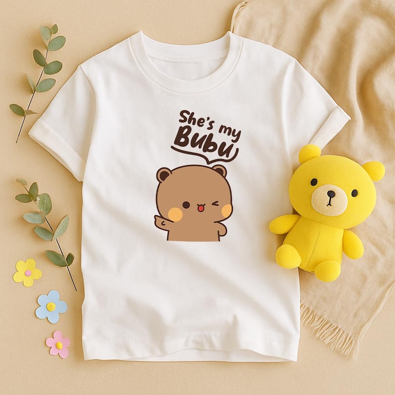 May include: White t-shirt featuring a cartoon bear with the text "She's my Bubu". A yellow teddy bear is next to the shirt. The shirt has short sleeves and a round neckline. The bear illustration is brown with pink cheeks.