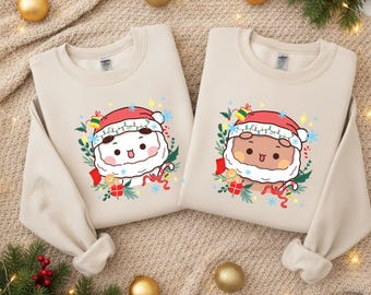 May include: Two beige sweatshirts with matching Santa Claus graphics. Each sweatshirt features a cartoon Santa face wearing a red hat, surrounded by festive elements like snowflakes, gifts, and greenery. The sweatshirts are perfect for the holiday season.