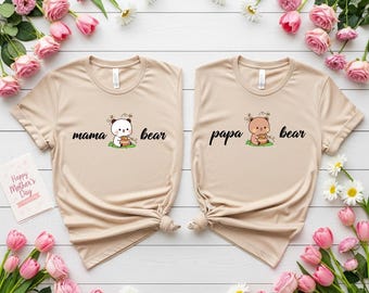 May include: Two beige t-shirts with the words "mama bear" and "papa bear" printed on them. Each shirt features a cartoon bear illustration. The shirts are surrounded by pink tulips, daisies, and greenery, with a "Happy Mother's Day" card.
