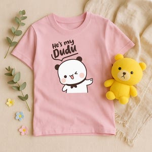 May include: A light pink t-shirt featuring a cartoon panda with the text "He's my Dudu". The panda has black ears, a black bow tie, and is winking. A yellow teddy bear is next to the shirt.