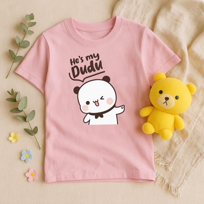 May include: A light pink t-shirt featuring a cartoon panda with the text "He's my Dudu". The panda has black ears, a black bow tie, and is winking. A yellow teddy bear is next to the shirt.