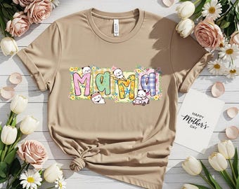 May include: A tan t-shirt with the word "Mama" in colorful, whimsical lettering. The letters are decorated with floral patterns and cute cartoon characters. A card with "Happy Mother's Day" is also visible.