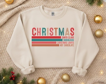 May include: A cream-colored sweatshirt with the word "CHRISTMAS" in a retro font. Below the word are three horizontal stripes in red, teal, and orange. The text "SANTA CLAUS, BUBU&DUDU, CHRISTMAS LIGHTS, HOT CHOCOLATE" is printed below the stripes.