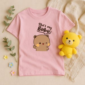 May include: A light pink t-shirt featuring a cartoon bear illustration with the text "She's my Bubu". A yellow teddy bear is next to the shirt. The shirt is made of soft material.