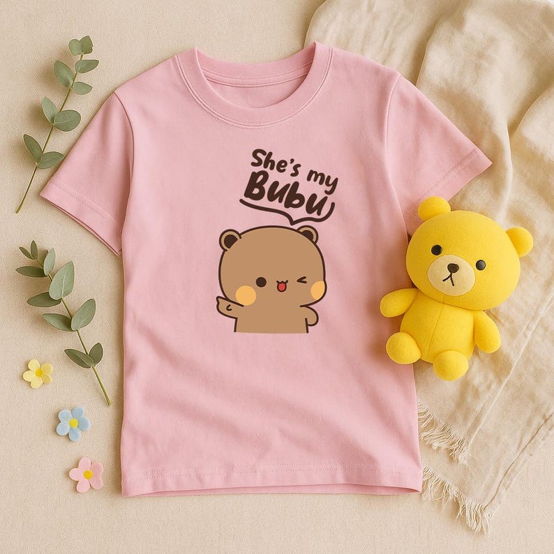 May include: A light pink t-shirt featuring a cartoon bear illustration with the text "She's my Bubu". A yellow teddy bear is next to the shirt. The shirt is made of soft material.