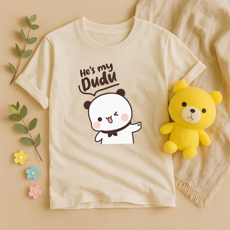 May include: Cream-colored t-shirt featuring a cartoon panda with the text "He's my Dudu." The panda is white with black accents and a bow tie. A yellow teddy bear is next to the shirt.