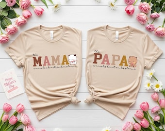 May include: Two beige t-shirts with the words "MAMA" and "PAPA" in brown and tan lettering. Each shirt has a small cartoon bear illustration and the phrase "wears her heart on her sleeve" or "wears his heart on his sleeve". The shirts are surrounded by pink roses and daisies.