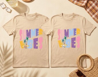 May include: Two beige t-shirts with the words "SUMMER VIBES" in colorful letters. The design includes palm trees and a cartoon character wearing a hat. The shirts are displayed with a drink, lemon slices, and a straw bag.