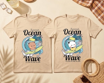 May include: Two beige t-shirts with a cartoon bear surfing on a wave, with the words "Summer Ocean Wave" printed above and below the image. The text "Life is better when you surf" is printed below the word "Wave".