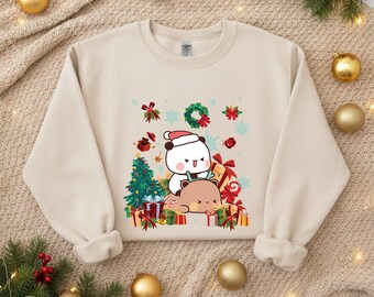 May include: A beige sweatshirt featuring a festive graphic of two cartoon bears surrounded by Christmas presents, a Christmas tree, and snowflakes. One bear wears a Santa hat. The design includes red, green, and white accents, perfect for the holiday season.