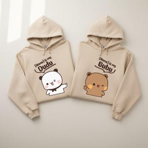 May include: Two beige hooded sweatshirts featuring cartoon bear graphics. The left hoodie displays a white bear with the text [Name] is my Dudu. The right hoodie shows a brown bear with the text [Name] is my Bubu. Both have long sleeves and drawstrings.