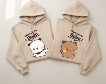 May include: Two beige hooded sweatshirts featuring cartoon bear graphics. The left hoodie displays a white bear with the text [Name] is my Dudu. The right hoodie shows a brown bear with the text [Name] is my Bubu. Both have long sleeves and drawstrings.