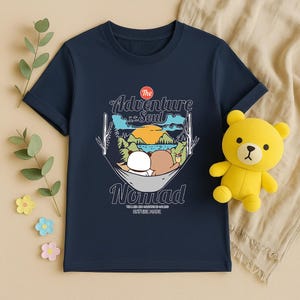 Peut inclure: T-shirt bleu marine avec un motif de deux personnages de dessins anim&eacute;s se relaxant dans un hamac, avec le texte "The Adventure Soul Nomad". Un ours en peluche jaune est plac&eacute; &agrave; c&ocirc;t&eacute; du t-shirt.