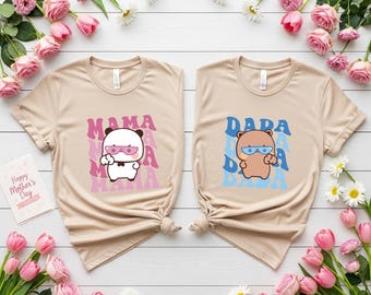 May include: Two beige t-shirts with cartoon graphics. One shirt features a white panda wearing pink sunglasses with the text "MAMA" in pink. The other shirt has a brown bear wearing blue glasses with the text "DADA" in blue.