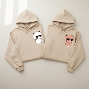 May include: Two beige hoodies with matching designs. One features a panda with sunglasses holding a phone, the other a bear with sunglasses holding a phone. Both hoodies have drawstrings and a kangaroo pocket.