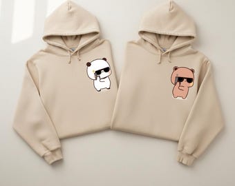 May include: Two beige hoodies with matching designs. One features a panda with sunglasses holding a phone, the other a bear with sunglasses holding a phone. Both hoodies have drawstrings and a kangaroo pocket.