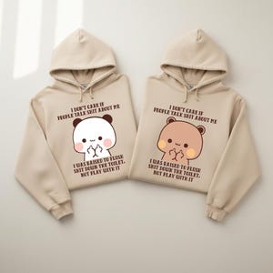 May include: Two beige hoodies with matching designs. Each hoodie features a cartoon animal face with a middle finger gesture and the text "I DON'T CARE IF PEOPLE TALK SHIT ABOUT ME" and "I WAS RAISED TO FLUSH SHIT DOWN THE TOILET, NOT PLAY WITH IT."