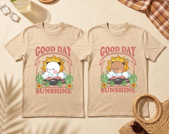 May include: Two beige t-shirts with the text "GOOD DAY SUNSHINE" in pink script. Each shirt features a cartoon bear wearing a sun hat, cacti, and sunflowers. The shirts are short-sleeved.