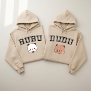 May include: Two beige hoodies with matching designs. One hoodie features the word "BUBU" above a white panda bear illustration. The other hoodie has the word "DUDU" above a brown bear illustration. Both hoodies have a drawstring hood.