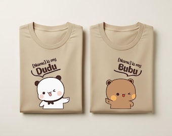 May include: Two beige t-shirts featuring cartoon bear graphics. The left shirt shows a white bear with the text [Name] is my Dudu. The right shirt shows a brown bear with the text [Name] is my Bubu. Both shirts have a round neckline and short sleeves.