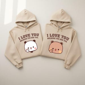 May include: Two beige hoodies with the text "I LOVE YOU BECAUSE YOU ARE YOU." Each hoodie features a cartoon animal graphic. One has a panda, the other a bear. Both are sticking out their tongues.