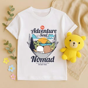 Peut inclure: T-shirt blanc avec un motif de deux ours de dessin anim&eacute; dans un hamac, avec les mots "Adventure Soul Nomad". Le motif comprend une sc&egrave;ne de montagne et de lac avec le texte "The lake and mountain is calling nature made". Un ours en peluche jaune est &agrave; c&ocirc;t&eacute; du t-shirt.