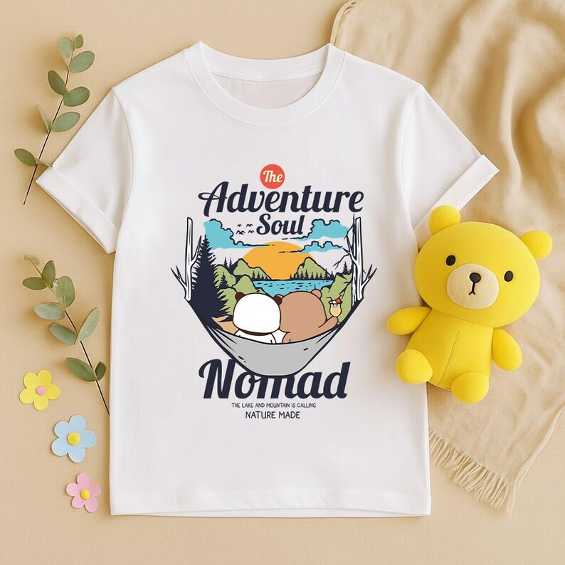 Peut inclure: T-shirt blanc avec un motif de deux ours de dessin anim&eacute; dans un hamac, avec les mots "Adventure Soul Nomad". Le motif comprend une sc&egrave;ne de montagne et de lac avec le texte "The lake and mountain is calling nature made". Un ours en peluche jaune est &agrave; c&ocirc;t&eacute; du t-shirt.