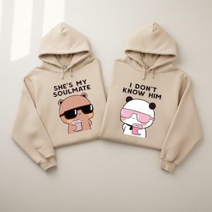 May include: Two beige hoodies with matching cartoon bear graphics. One hoodie says "SHE'S MY SOULMATE" and the other says "I DON'T KNOW HIM". The bear graphics wear sunglasses and hold coffee cups. The hoodies have drawstrings.