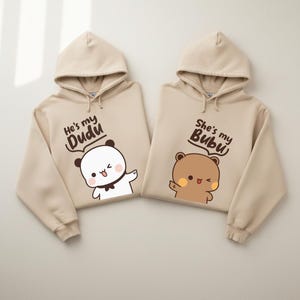 Puede incluir: Dos sudaderas con capucha beige con gráficos de osos y pandas de dibujos animados a juego. Una sudadera dice "He's my Dudu" con un panda blanco. La otra dice "She's my Bubu" con un oso pardo.