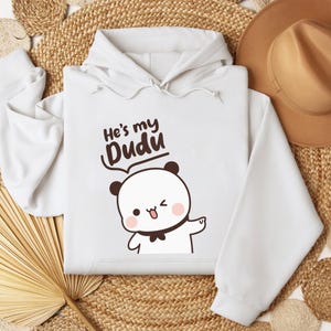 May include: White hoodie with the text "He's my Dudu" above a cartoon panda bear winking and sticking out its tongue. The panda has black ears, a black bow tie, and pink cheeks. The hoodie has a drawstring hood.