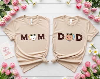 May include: Two beige t-shirts with the words "MOM" and "DAD" printed in brown. Each word has a cartoon character with sunglasses. The shirts are surrounded by pink roses, daisies, and greenery, with a "Happy Mother's Day" card.