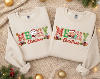 May include: Two beige sweatshirts with the words "MERRY Christmas" in green and red lettering. Each sweatshirt features a cartoon character wearing a Santa hat and holding a gift bag. Christmas-themed apparel.