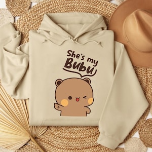 May include: A beige hoodie with a hood and long sleeves. The front features a cartoon bear illustration with the text "She's my Bubu" in a playful font. The bear is brown with orange cheeks and a small, red tongue.