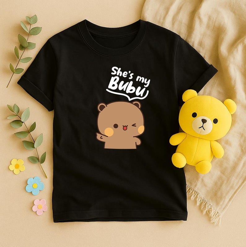 May include: Black t-shirt with the text "She's my Bubu" above a cartoon bear illustration. A yellow teddy bear plush toy is next to the shirt. The shirt is made of a soft material.