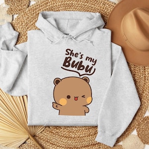 May include: A light gray hoodie with a cartoon bear illustration and the text "She's my Bubu". The bear is brown with orange cheeks and is winking. The hoodie has a drawstring hood and long sleeves.