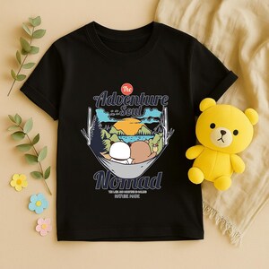 Peut inclure: T-shirt noir avec un motif de deux ours en peluche dans un hamac, avec le texte "The Adventure Soul Nomad". Un ours en peluche jaune est sur le c&ocirc;t&eacute; droit du t-shirt.