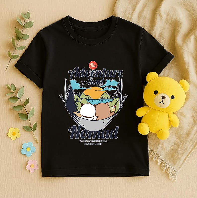 Peut inclure: T-shirt noir avec un motif de deux ours en peluche dans un hamac, avec le texte "The Adventure Soul Nomad". Un ours en peluche jaune est sur le c&ocirc;t&eacute; droit du t-shirt.