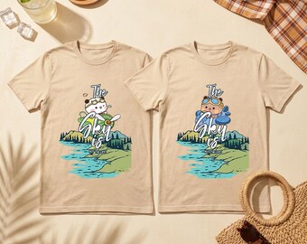 May include: Two beige t-shirts with a graphic of a bear wearing goggles and a pilot's hat, and a panda wearing a pilot's hat and goggles. The text "The Sky is" is below the characters. The graphic is above a landscape with water and mountains.