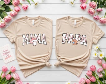 May include: Two beige t-shirts with the words "MAMA" and "PAPA" in a baseball-themed font. Each shirt features a cartoon character wearing a baseball cap. The shirts are surrounded by pink flowers and greenery.