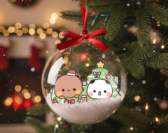 May include: A clear glass Christmas ornament with a red ribbon, hanging on a Christmas tree. Inside the ornament are two cartoon characters, a brown bear and a panda, with a Christmas tree and snow.