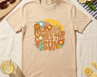 May include: Beige t-shirt with a retro design featuring the text "Here Comes The Sun" surrounded by flowers and a sun graphic. The shirt is displayed with sunglasses, a straw hat, and a drink with lemon slices.