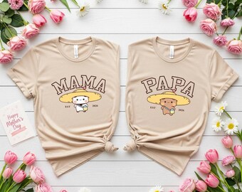 May include: Two beige t-shirts with the words "MAMA" and "PAPA" printed on them in brown. Each shirt features a cartoon character wearing a yellow hat. The shirts are surrounded by pink flowers and greenery.