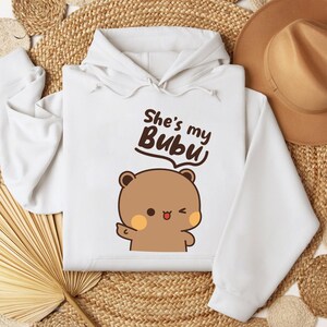 May include: White hoodie with the text "She's my Bubu" above a cartoon bear with a wink and a raised paw. The bear is brown with peach-colored cheeks. The hoodie has a drawstring hood.