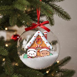 May include: A clear glass Christmas ornament with a red ribbon. Inside, a gingerbread house with snow-covered roof and two cartoon characters wearing Santa hats. The ornament hangs on a green Christmas tree.