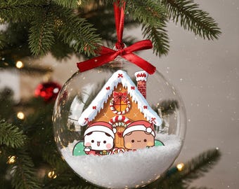 May include: A clear glass Christmas ornament with a red ribbon. Inside, a gingerbread house with snow-covered roof and two cartoon characters wearing Santa hats. The ornament hangs on a green Christmas tree.