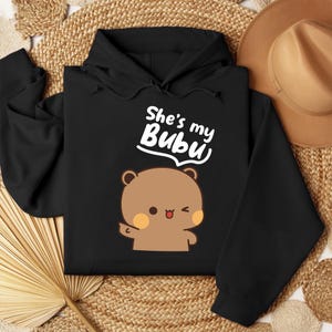 May include: Black hoodie featuring a cartoon bear with a wink and the text "She's my Bubu" in white. The bear is brown with peach-colored cheeks. The hoodie has a drawstring hood and long sleeves.